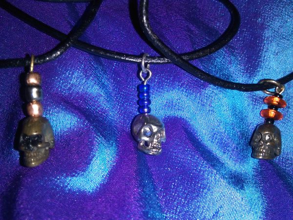 Skull necklaces on 9" black cord: (1) brass skull with pearl & silver beads; (1) brass skull with orange & black beads; (1) pewter skull with blue beads - $25