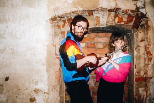 A photo of two people wearing brightly coloured windbreakers in a brick alcove, holding a violin and a flute