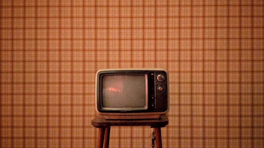 Un vecchio televisore poggiato sopra un tavolino di legno davanti a una parete quadrettata conduce ai video di Giacomo Deiana.