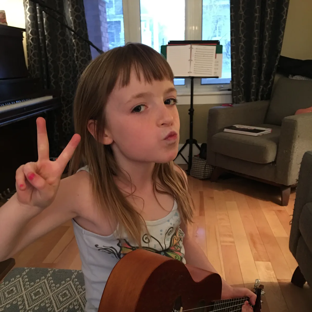 Young girl sitting and holding a ukulele while giving a "peace sign"