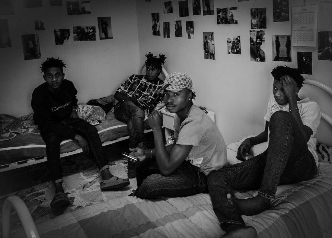 I: Nigerian Youth Migrants at Camp - April 2016, Syracuse, Sicily.  A group of young migrants from Nigeria spend a weekday in the bedroom of their camp, a six-bedroom apartment that houses 15 unaccompanied West African youth. There are around 20 small-scale youth migrant camps in Syracuse, Sicily which house migrants of similar geographical and linguistic backgrounds. The migrants pictured speak English, and the other rooms in the camp are inhabited by either French- or English-speaking West Africans. Residents of this camp spend the vast majority of their days in the camp surfing the internet and listening to music, or in the common room socializing, playing games, cooking and eating.

