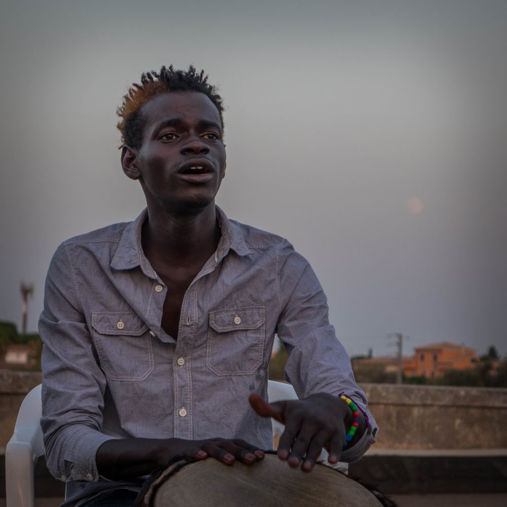 Ibrahim Sanneh – August 2017, Syracuse, Sicily. Ibrahim, a 17-year-old migrant from The Gambia, sings and plays a drum in preparation for a concert the following night at Lido Terraza Fanusa, a nearby beach club.