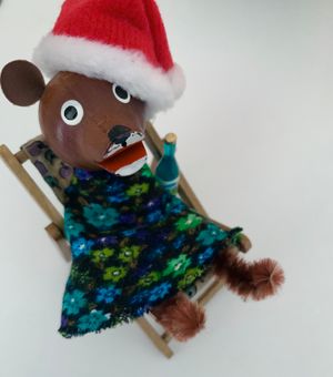Bear wooden finger puppet sat in chair wearing a Christmas hat, with a bottle of Blue Nun.