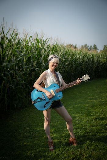Woman standing while holding a blue guitar, outdoor setting..

