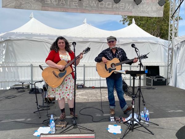 Image Description: A person with curly brown hair in a dress with an apple print and a red sweater and a man with a black Western shirt and jeans standing on a stage.  Both are playing guitars.