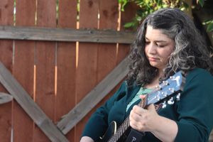 Image Description: A nonbinary person with curly brown hair (with some white streaks) playing guitar.