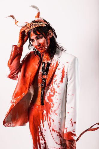 Medusa, trans nonbinary alt-pop musician and rapper; white suit, tuxedo, tiara, horns; bouquet of flowers; fake blood; harness; bare chest; prom scene, Carrie; photographer Justin Ruggiero