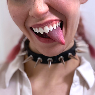 Nose, mouth, hair, and neck of Medusa, non-binary musician; pop artist; rapper; hip-hop artist; pink hair in pigtail braids, tongue stuck out under teeth, both pointed; smiling; spike collar