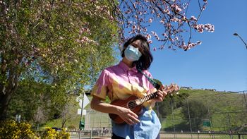 nonbinary person holding a ukulele with a pineapple strap in front of a cherry tree
