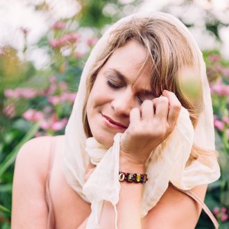 Anic Asha (Anic Proulx), meditation music singer in a relaxation pose with a scarf on her head , eyes closed and hand touching her face. Pink purple gentle flowers in the background.