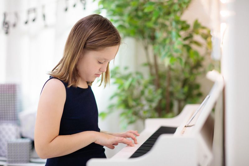 Une enfant jouant du piano avec un air concentré regardant le piano et une plante en arrière. Décor doux avec piano blanc.