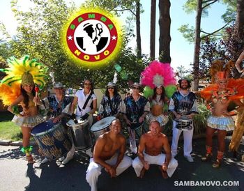 Six samba drummers, two samba dancers and two capoeira performers, all posing outside at a country club on a bright sunny day
