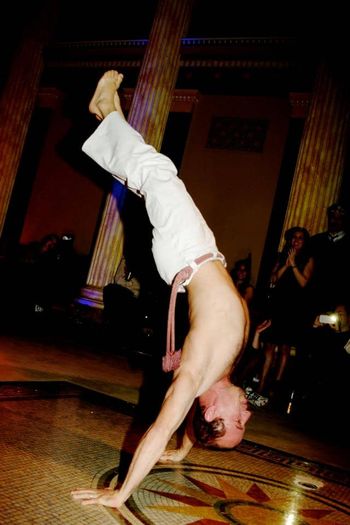 A capoeira party entertainer in great shape with his shirt off Standing on both hands while a crowd of event guests watches
