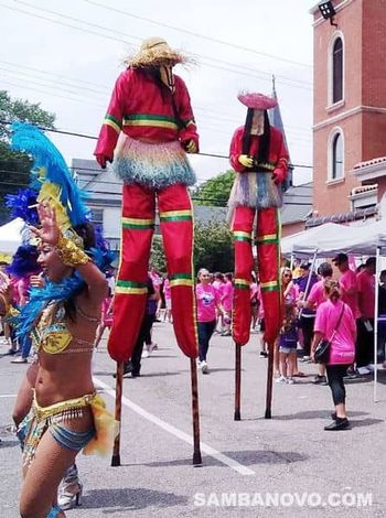 Two giant stilt walkers called Moko Jumbies, walking in a parade at a street fair with samba dancers while people watch
