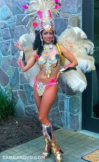 This is a samba dancer for hire in NYC posing outside before in a pink & silver costume covered with feathers and sequins

