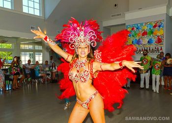 Dancer wearing a red costume dancing to live samba drummers at a party indoors with a large crowd watching

