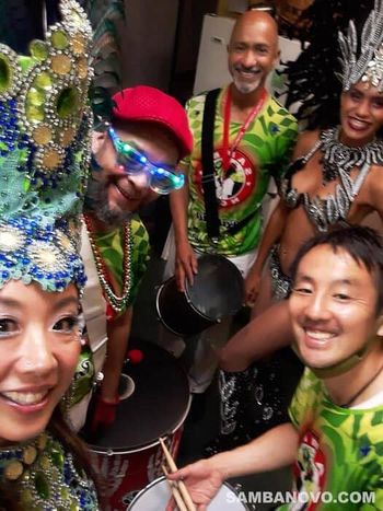A smiling group of 5 Brazilian samba dancers & drummers in bright green and red costumes after giving a custom samba show
