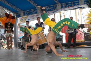 Two Capoeira performers on stage, on their heads, giving a performance with samba drummers playing live behind them
