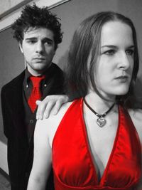 A black and white photo of a white man with short dark hair and a woman with long dark hair - he has his hand on her shoulder and the only colorful items are his red tie and her red dress